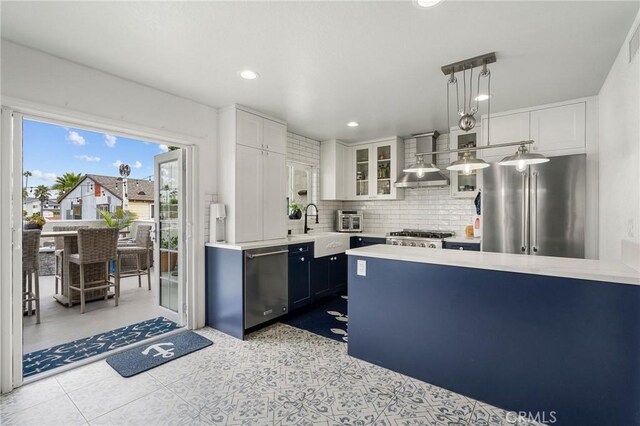 kitchen with appliances with stainless steel finishes, white cabinetry, hanging light fixtures, and wall chimney range hood