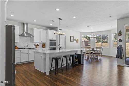 kitchen with dark wood-type flooring, a kitchen island with sink, white cabinets, wall chimney exhaust hood, and decorative light fixtures