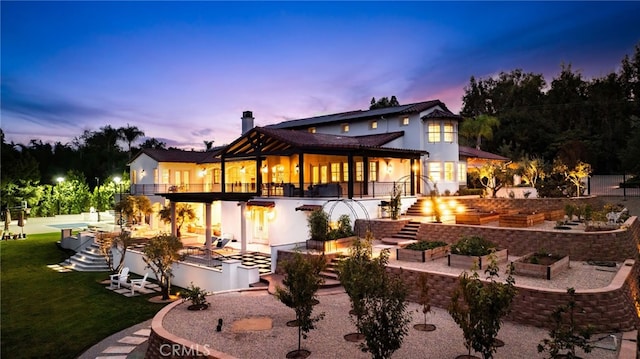 back house at dusk with a balcony, a patio, and a lawn