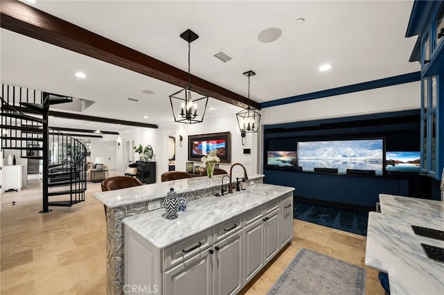 kitchen with gray cabinetry, light stone countertops, sink, an island with sink, and beamed ceiling