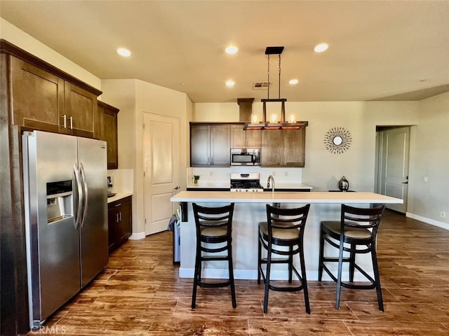 kitchen with hanging light fixtures, dark hardwood / wood-style flooring, stainless steel appliances, dark brown cabinets, and a center island with sink