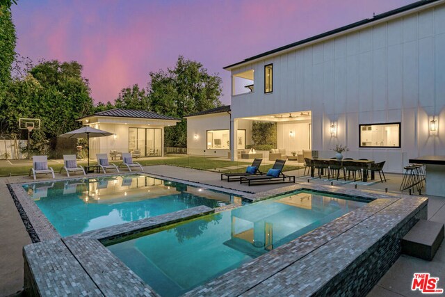 pool at dusk with a patio area, an in ground hot tub, a yard, and an outdoor structure