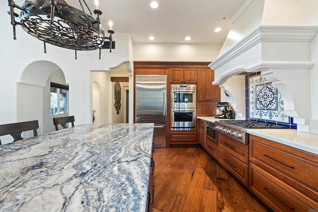 kitchen featuring dark wood-type flooring, a breakfast bar area, backsplash, built in appliances, and light stone counters