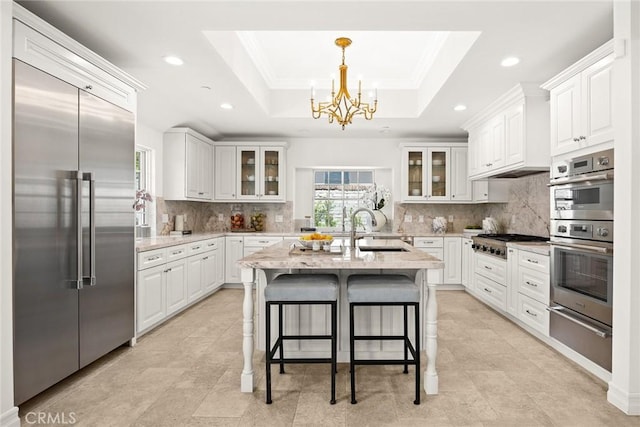 kitchen with stainless steel appliances, a tray ceiling, a kitchen island with sink, sink, and white cabinetry