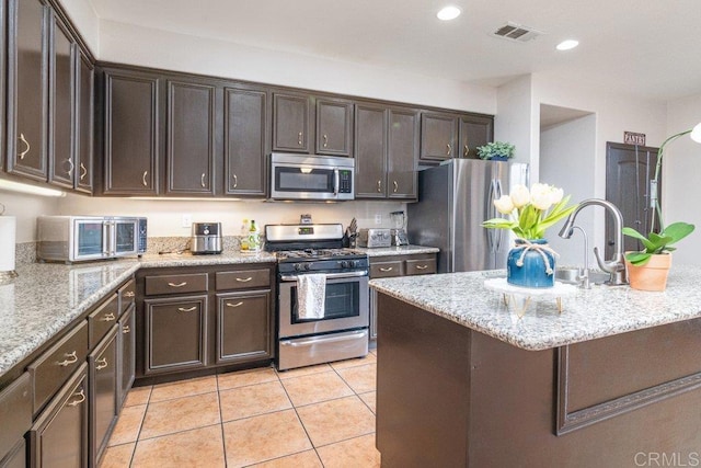 kitchen featuring dark brown cabinetry, light stone countertops, light tile patterned flooring, and appliances with stainless steel finishes