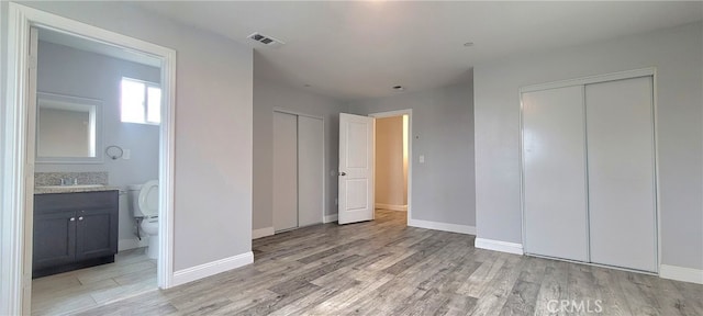 unfurnished bedroom with two closets, light wood-type flooring, ensuite bath, and sink