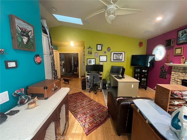 home office featuring ceiling fan, light wood-type flooring, a stone fireplace, and vaulted ceiling with skylight