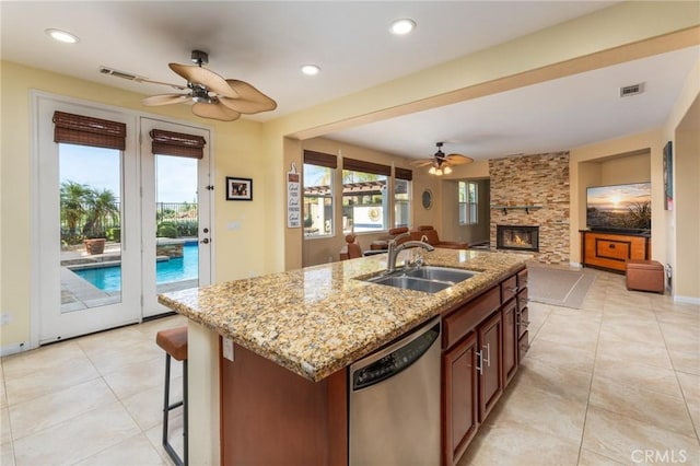 kitchen featuring light stone countertops, dishwasher, sink, a stone fireplace, and a center island with sink