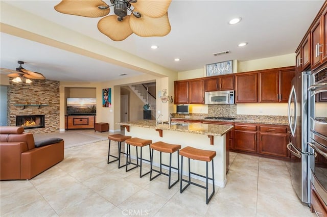 kitchen featuring light stone countertops, stainless steel appliances, a stone fireplace, a kitchen island with sink, and a breakfast bar