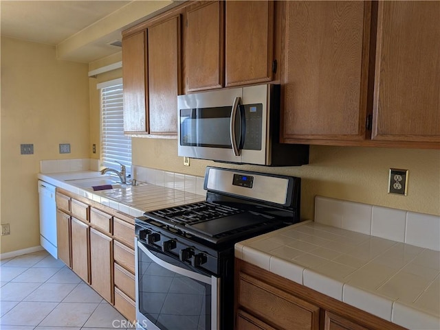 kitchen with tile countertops, appliances with stainless steel finishes, brown cabinetry, light tile patterned flooring, and a sink