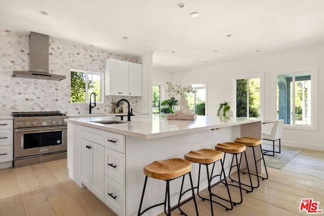 kitchen featuring stainless steel range, an island with sink, wall chimney range hood, and a healthy amount of sunlight