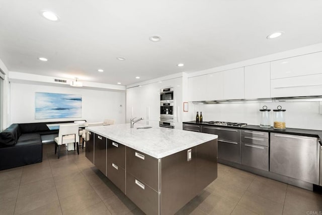 kitchen with light stone countertops, sink, white cabinetry, stainless steel gas stovetop, and an island with sink