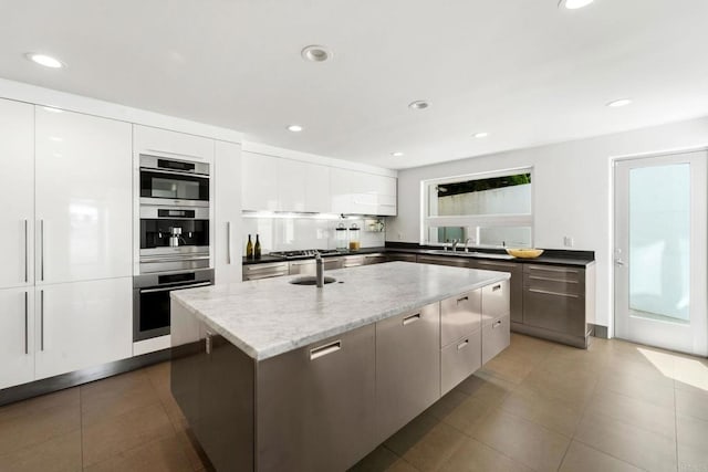 kitchen with sink, light stone counters, white cabinetry, and an island with sink