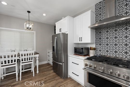 kitchen featuring wall chimney range hood, hanging light fixtures, dark wood-type flooring, appliances with stainless steel finishes, and white cabinets