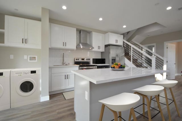 kitchen with wall chimney range hood, appliances with stainless steel finishes, white cabinetry, washing machine and clothes dryer, and a kitchen bar