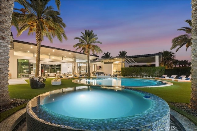 pool at dusk featuring a lawn and a hot tub