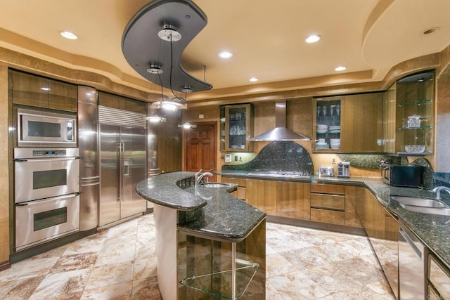 kitchen featuring sink, built in appliances, dark stone counters, hanging light fixtures, and wall chimney exhaust hood