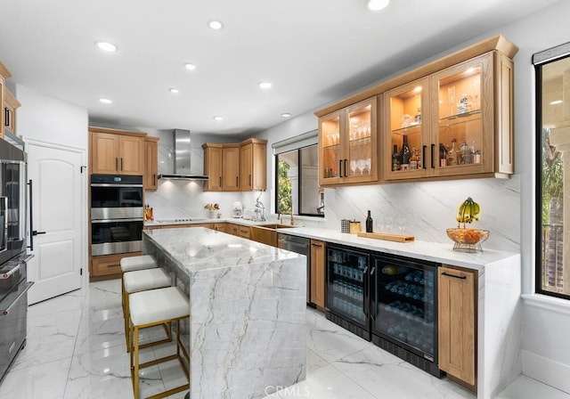 kitchen with light stone counters, beverage cooler, wall chimney range hood, appliances with stainless steel finishes, and a kitchen bar