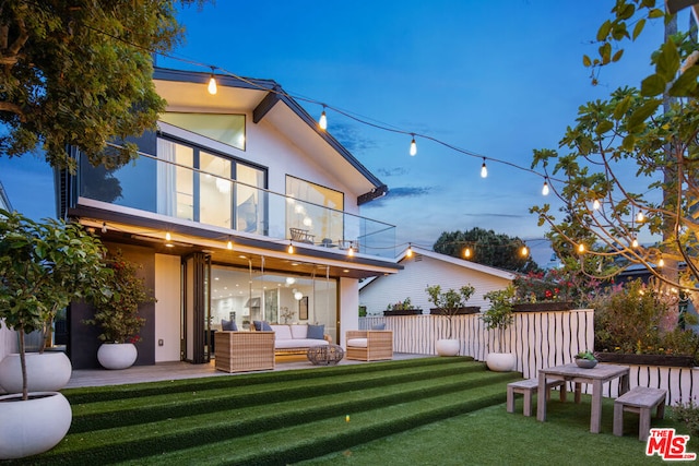 back house at dusk featuring a lawn, a patio area, a balcony, and an outdoor hangout area