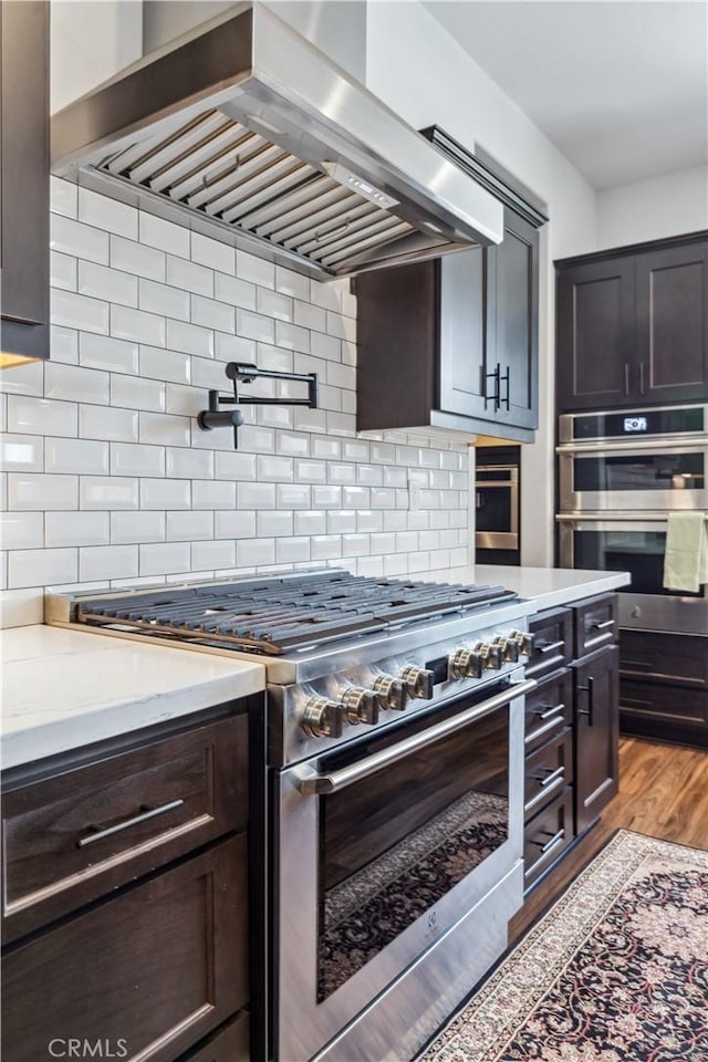 kitchen featuring dark brown cabinetry, light hardwood / wood-style flooring, stainless steel appliances, range hood, and decorative backsplash