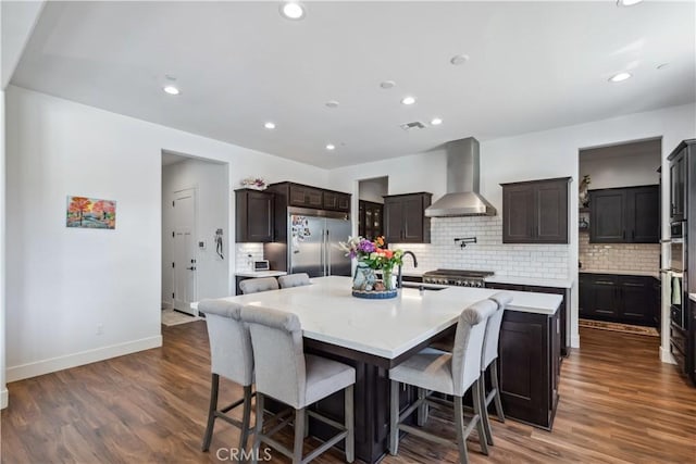 kitchen featuring appliances with stainless steel finishes, a breakfast bar, decorative backsplash, a kitchen island with sink, and wall chimney range hood