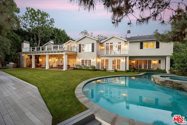 back house at dusk featuring a balcony, a patio, a pool with hot tub, and a yard