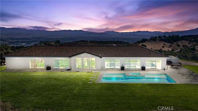 back house at dusk featuring a mountain view, a pool with hot tub, a patio area, and a lawn