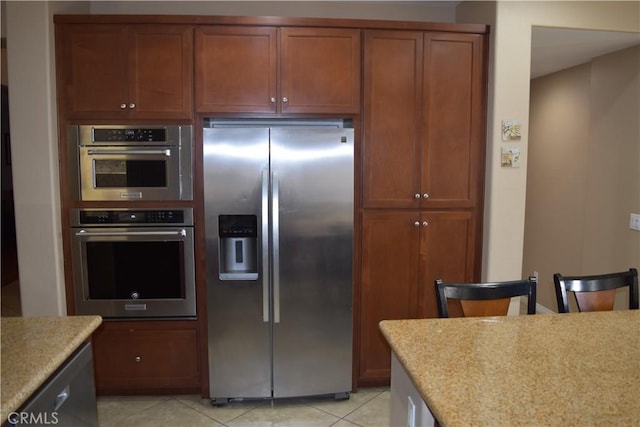 kitchen featuring light tile patterned floors, appliances with stainless steel finishes, brown cabinetry, and light stone countertops