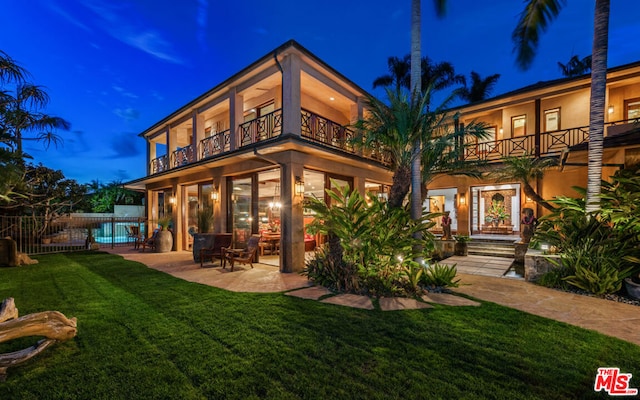 back house at twilight with a balcony, a yard, and a patio