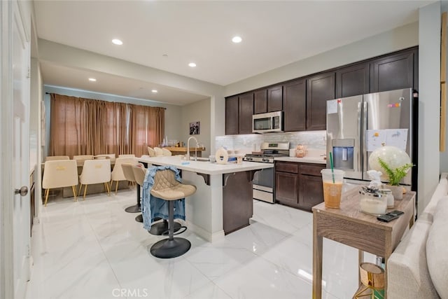 kitchen featuring decorative backsplash, appliances with stainless steel finishes, a kitchen breakfast bar, a kitchen island with sink, and dark brown cabinetry