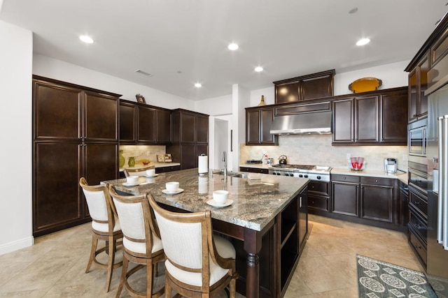 kitchen with backsplash, light stone countertops, exhaust hood, dark brown cabinetry, and a kitchen island with sink