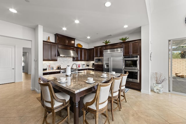 kitchen with a breakfast bar area, built in appliances, exhaust hood, stone counters, and a kitchen island with sink