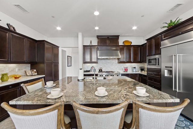 kitchen with an island with sink, light stone counters, built in appliances, and a breakfast bar area