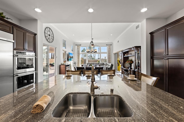 kitchen with appliances with stainless steel finishes, dark brown cabinets, an inviting chandelier, decorative light fixtures, and dark stone counters