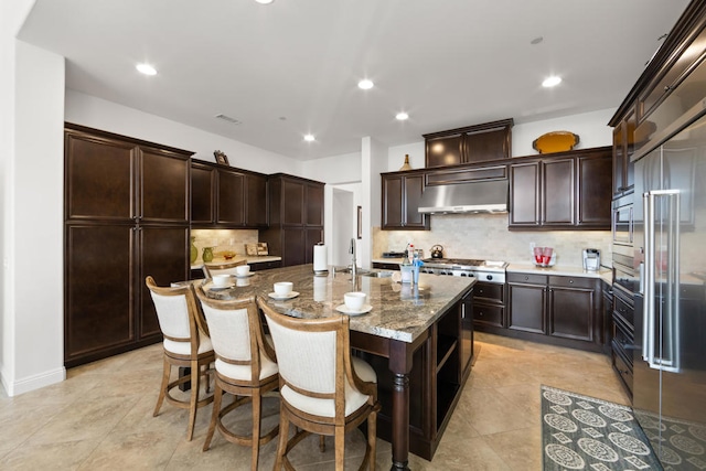 kitchen with dark brown cabinetry, an island with sink, backsplash, range hood, and light stone countertops