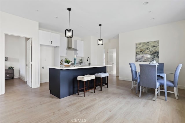 kitchen featuring pendant lighting, an island with sink, light hardwood / wood-style flooring, wall chimney range hood, and white cabinetry