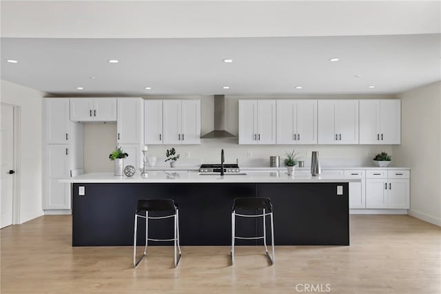 kitchen with white cabinets, a kitchen island with sink, wall chimney exhaust hood, and light wood-type flooring