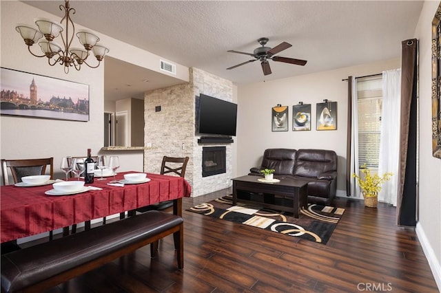 dining space featuring dark wood-type flooring, ceiling fan with notable chandelier, a stone fireplace, and a textured ceiling