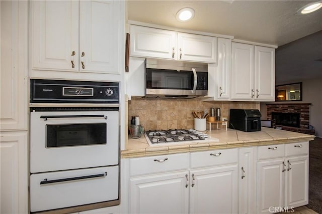 kitchen featuring white cabinetry, tile counters, a brick fireplace, backsplash, and white appliances