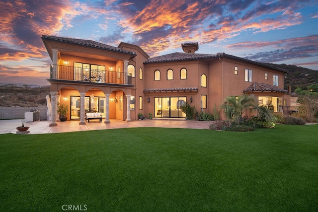 back house at dusk featuring a lawn, a balcony, and a patio area