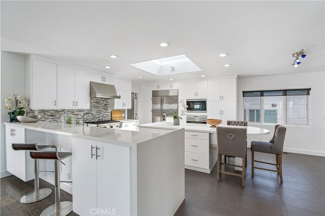 kitchen with white cabinets, a breakfast bar, built in appliances, ventilation hood, and a skylight