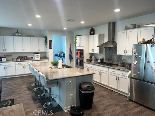 kitchen with wall chimney exhaust hood, stainless steel appliances, dark wood-type flooring, a center island with sink, and white cabinetry