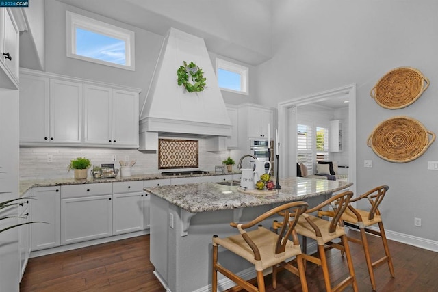 kitchen with premium range hood, a high ceiling, a center island with sink, sink, and light stone countertops