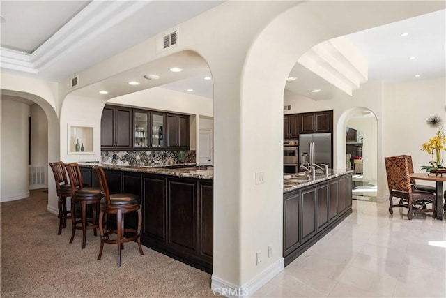 bar with dark stone countertops, sink, backsplash, stainless steel fridge, and dark brown cabinets