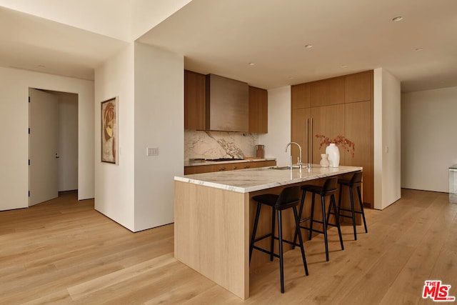 kitchen with sink, a center island with sink, light wood-type flooring, a kitchen bar, and decorative backsplash