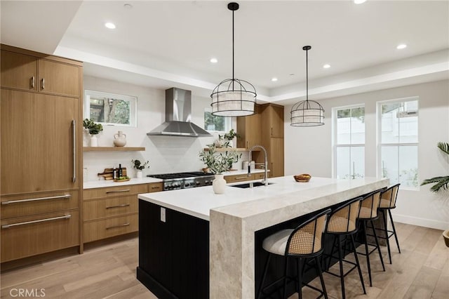 kitchen with a raised ceiling, stove, light wood-style floors, a sink, and wall chimney range hood