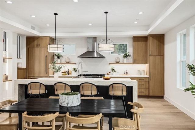 kitchen featuring light wood-style floors, wall chimney range hood, open shelves, brown cabinetry, and a center island with sink