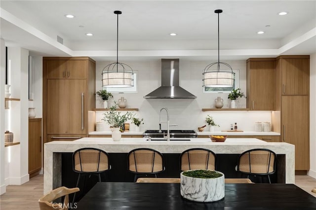 kitchen featuring a breakfast bar, light wood-style floors, wall chimney range hood, brown cabinets, and open shelves