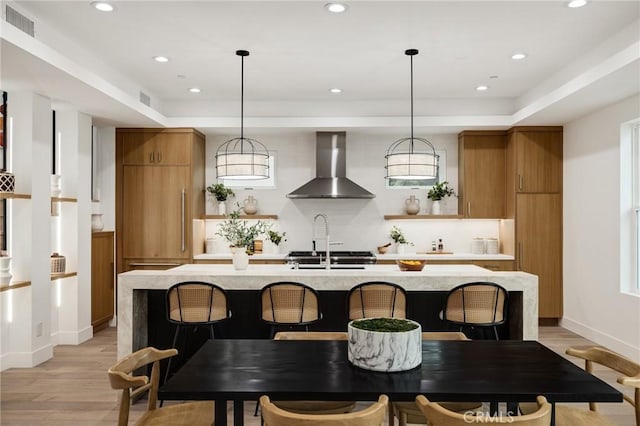 kitchen with visible vents, light wood-style floors, brown cabinets, wall chimney exhaust hood, and a center island with sink