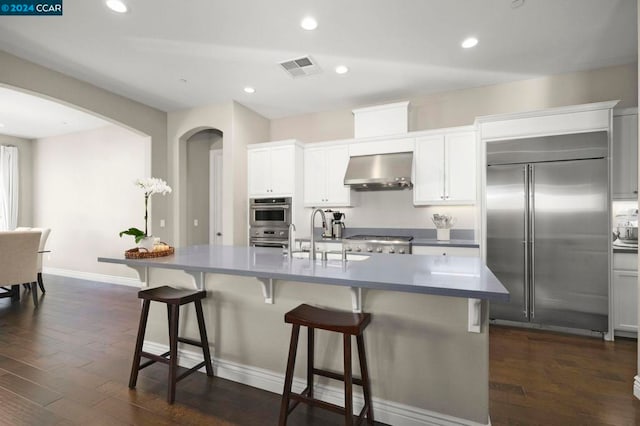 kitchen featuring appliances with stainless steel finishes, sink, wall chimney exhaust hood, dark wood-type flooring, and a center island with sink